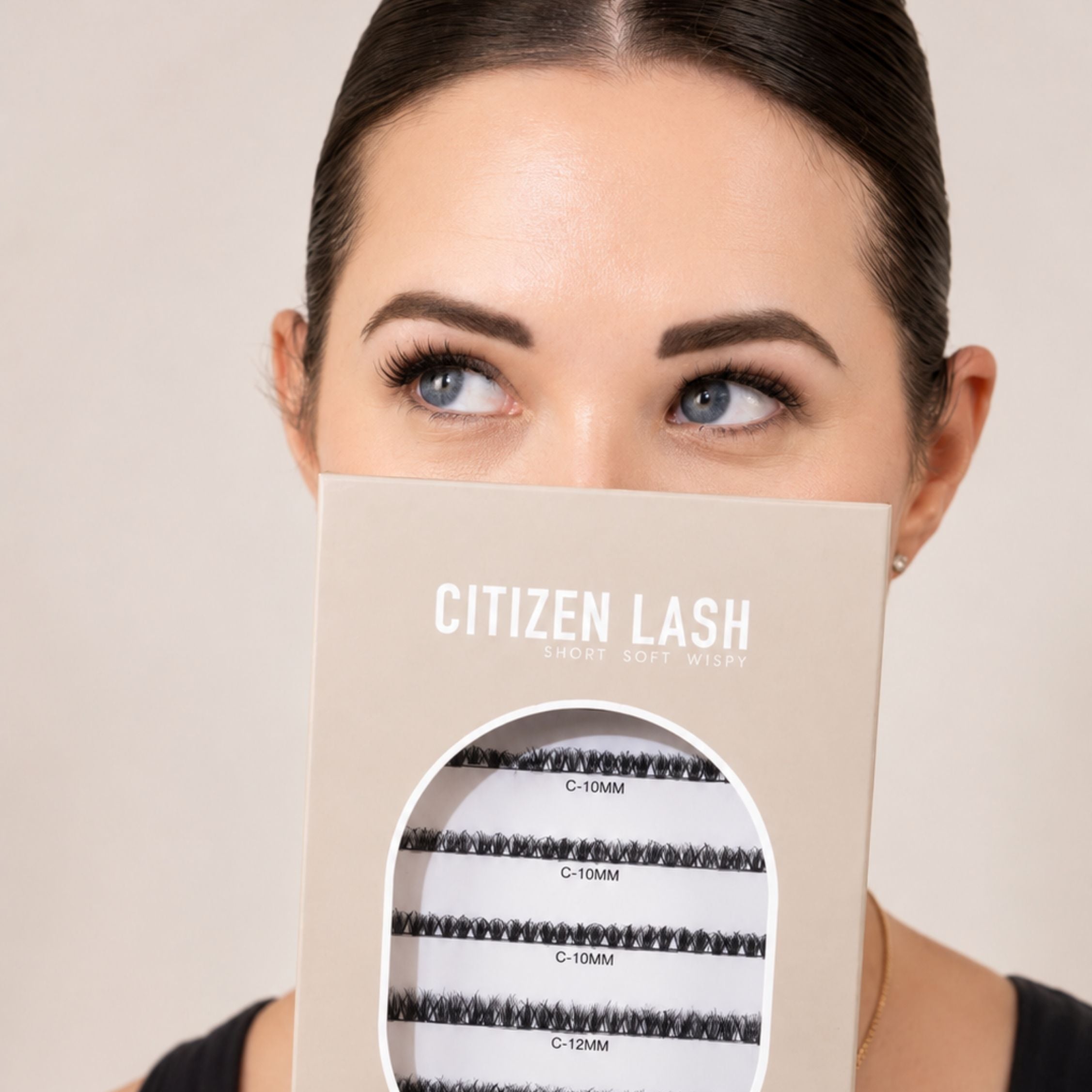 A woman holds a beige box of Citizen Lash false eyelashes in front of her face. The box features a clear oval window displaying black lash clusters labeled with different lengths (C-10MM, C-10MM, C-10MM, C-12MM).